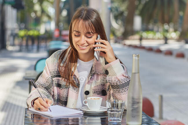 Positive young woman taking notes in notebook during phone conversation while sitting at table in outdoor cafe in city — Stock Photo