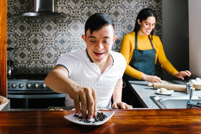 Encantado adolescente étnico com síndrome de Down decorar biscoitos com chips de chocolate enquanto prepara pastelaria com mãe sorridente em casa — Fotografia de Stock