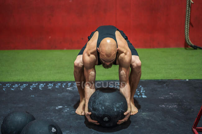 Сильный спортсмен поднимает тяжелый мяч с пола во время тренировки в современном спортзале — стоковое фото