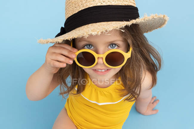 Ângulo alto de linda menina feliz vestindo maiô amarelo e chapéu de palha com óculos de sol elegantes sentados no fundo azul e olhando para a câmera — Fotografia de Stock