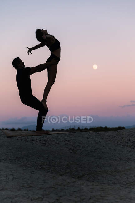 Вид сбоку силуэта неузнаваемой гибкой женщины, стоящей на ногах мужчины во время сеанса акро-йоги на фоне вечернего неба — стоковое фото