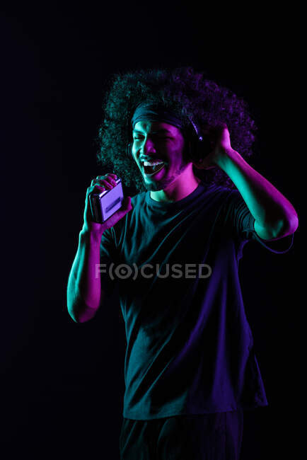 Carefree Latin male with vintage cassette player and in headphones listening to music and singing songs on black background in studio with neon lights — Stock Photo