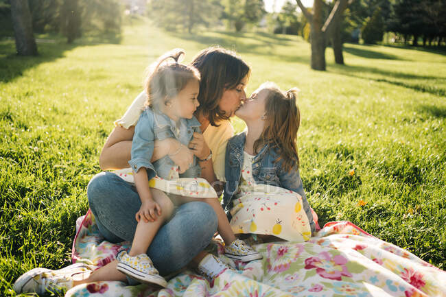Милые сестрички целуют счастливую мать, отдыхая вместе на одеяле на зеленой лужайке в летнем парке. — стоковое фото