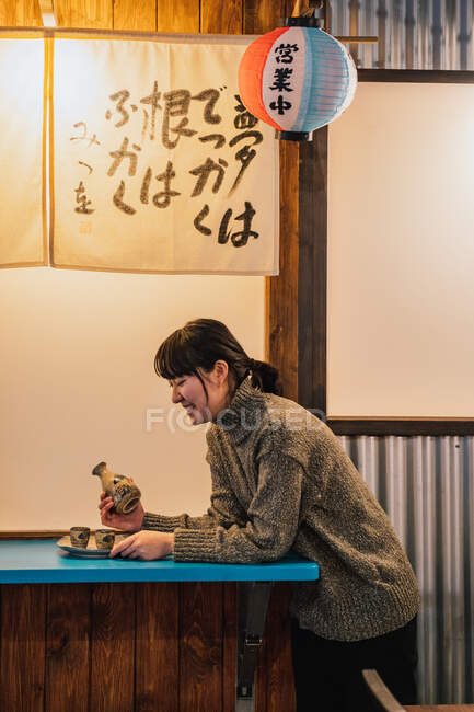 Vista lateral da mulher asiática exultante em suéter sentado à mesa azul com bebida tradicional no café — Fotografia de Stock