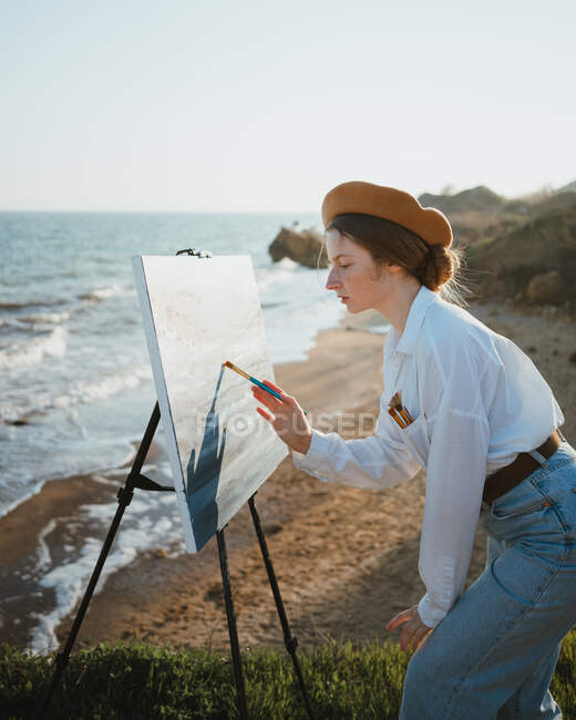 Вид сбоку на молодую женщину в стильной одежде и берете, стоящую на травянистом побережье возле песка и океана в солнечный день, рисуя картину кистью на холсте на мольберте — стоковое фото