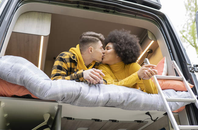 Side view of diverse man and woman kissing each other while lying down on bed in modern van during road trip — Stock Photo