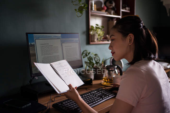 Side view of Asian female freelancer reading notes in notebook while sitting at table with computer and working on project at home — Stock Photo