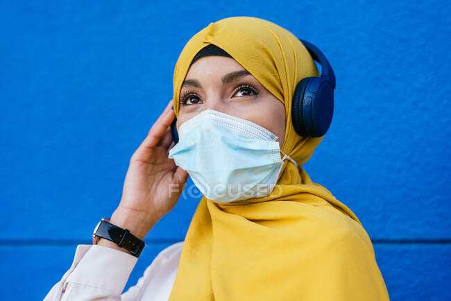 Mujer musulmana en hiyab y máscara protectora escuchando música en auriculares sobre fondo azul en la ciudad - foto de stock