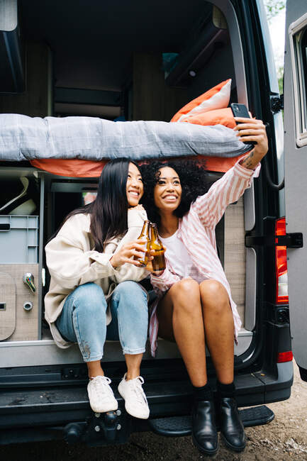 Corpo inteiro multirracial jovens mulheres clinking garrafas de cerveja e tomar selfie enquanto sentado em van durante a viagem de carro — Fotografia de Stock