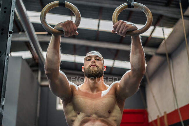 Strong shirtless man looking at camera standing doing exercise on gymnastic rings during intense workout in modern gym — Stock Photo