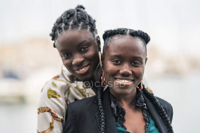 Задоволені стильними афроамериканськими жінками, які тримаються поруч і дивляться на камеру з задумливою посмішкою в парку в яскравий день. — стокове фото