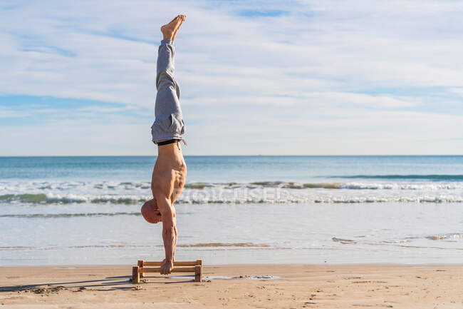 Vue latérale sur toute la longueur de l'athlète masculin exécutant handstand avec barres parallèles sur la côte sablonneuse avec des vagues de l'océan sur fond — Photo de stock