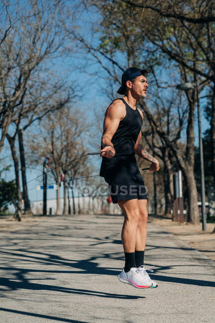 Male athlete in sportswear jumping with skipping rope and looking away on walkway during cardio training in park — Stock Photo