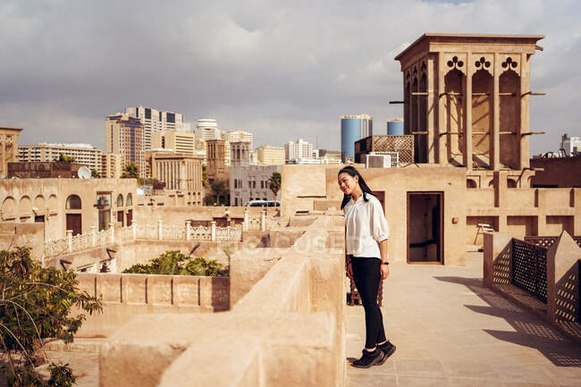 Vista lateral de la joven turista curiosa parada en la terraza de la casa de piedra envejecida durante el turismo en el Barrio Histórico Al Fahidi en Dubai - foto de stock