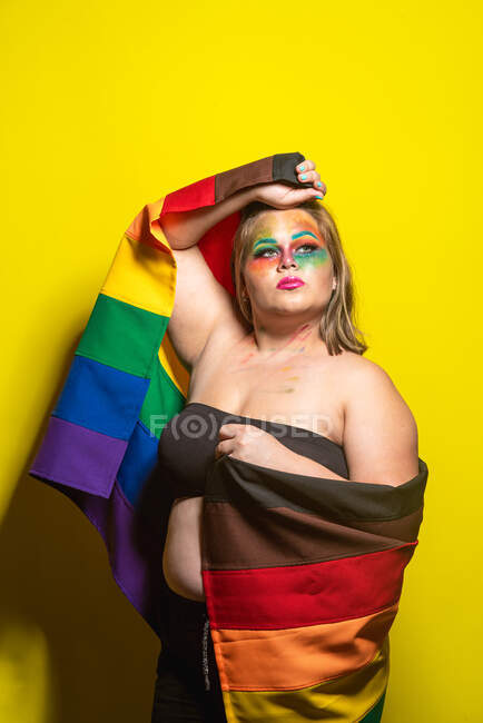 Надлишкова жіноча модель з креативним макіяжем, що показує прапор ЛГБТ і дивиться на жовте тло — стокове фото