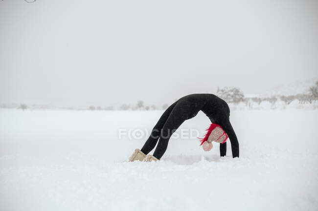Vista lateral de delgada hembra con pelo rosa haciendo yoga en Urdhva Dhanurasana en prado nevado de invierno - foto de stock