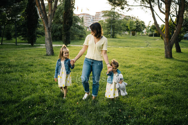 Full body of young woman holding hands of adorable little daughters in similar outfits while walking together on green grassy lawn in summer park — Stock Photo