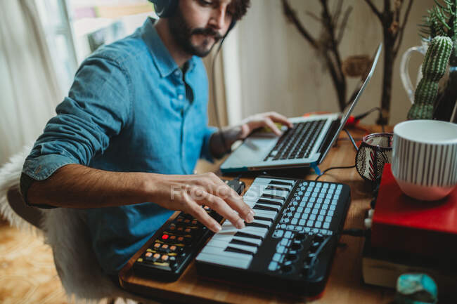 Seitenansicht eines jungen Mannes mit Kopfhörern, der Synthesizer und Laptop am heimischen Tisch benutzt — Stockfoto