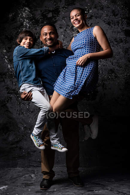 Entzückter ethnischer Mann hält Frau und Sohn und hat Spaß im Studio auf dunklem Hintergrund — Stockfoto