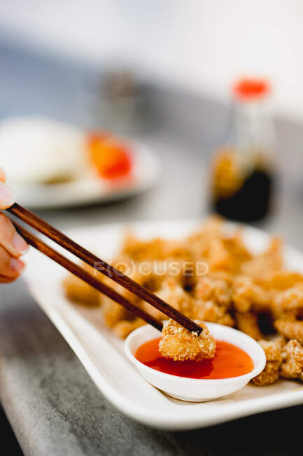 Рука неузнаваемой женщины, использующей палочки для еды, чтобы окунуть вкусный вонтон в соус на размытом фоне ресторана — стоковое фото