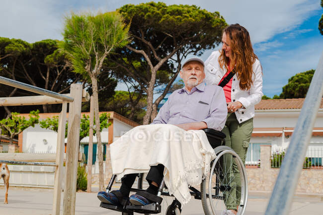Femme adulte souriante et homme âgé en fauteuil roulant se détendre ensemble en ville par une journée ensoleillée — Photo de stock