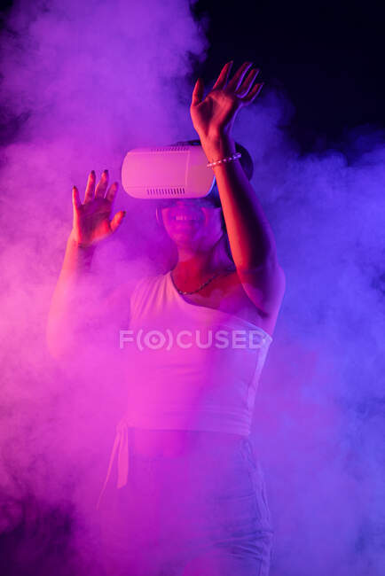 Невпізнавана жінка в гарнітурі VR, що взаємодіє з віртуальною реальністю, стоячи в темній студії з паровим і синім і рожевим неоновим освітленням — стокове фото