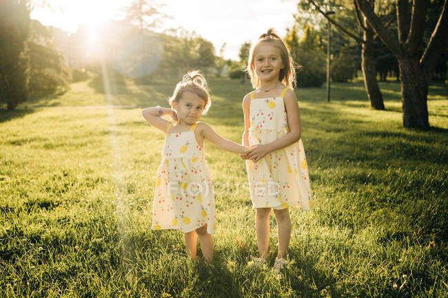 Cute toddler girl with older sister wearing similar sundresses holding hands and looking at camera while playing together on green grassy meadow in sunny summer park — Stock Photo