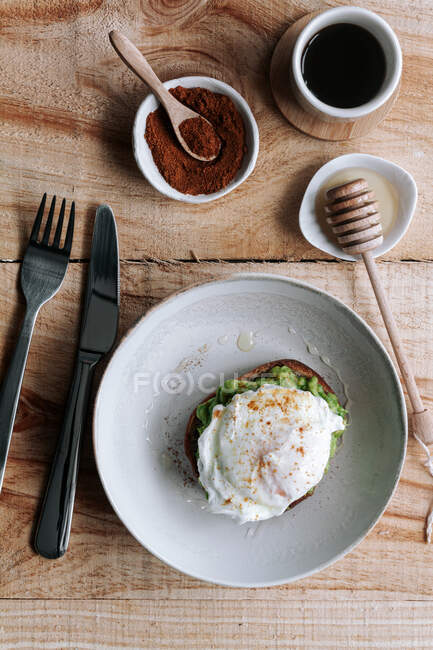 Apetitivo trozo de pan de grano con aguacate y huevo escalfado en la parte superior colocado en el plato en la mesa de madera - foto de stock
