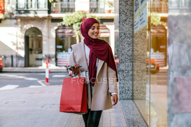 Deleitado comprador musulmán en pañuelo para la cabeza y con bolsas de compras de pie cerca de escaparate de la tienda en la ciudad - foto de stock