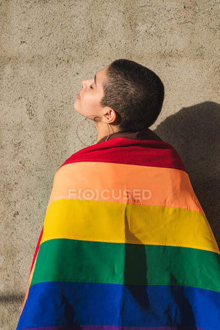 Серйозна молода бісексуальна етнічна жінка з різнокольоровим прапором символів ЛГБТК і закритими очима в сонячний день — стокове фото