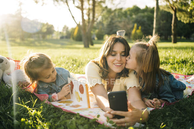 Happy young woman and cute little daughters lying on blanket and taking selfie on smartphone while having fun together on green meadow in summer park — Stock Photo
