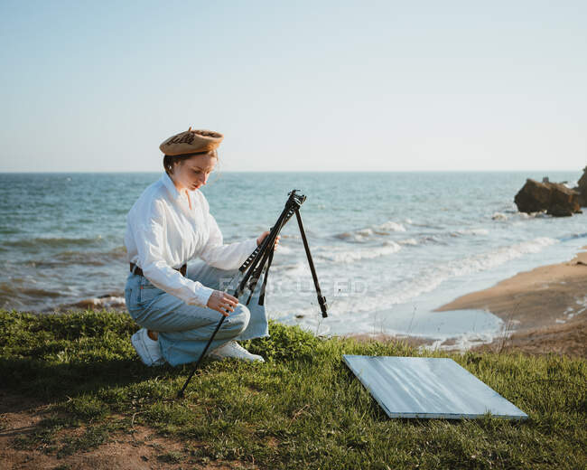 Side view of young stylish female painter in casual clothes and beret setting up easel on grassy terrain above sandy beach of wavy ocean on sunny day — Stock Photo