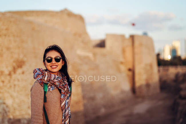 Cheerful young female traveler in sunglasses standing against stone wall of historic archaeological site Qal at al Bahrain or Bahrain Fort — Stock Photo
