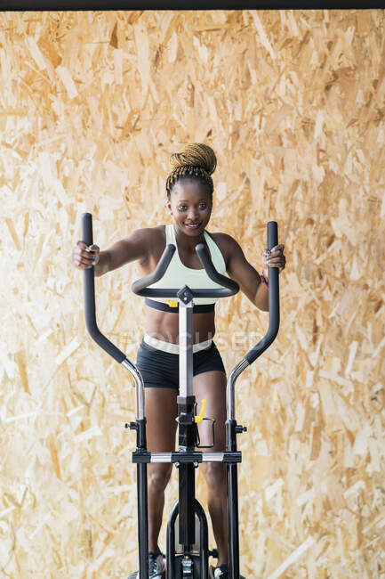 Joven atleta femenina afroamericana alegre en ropa deportiva haciendo ejercicio en la máquina de ciclismo mientras mira hacia otro lado en el gimnasio - foto de stock