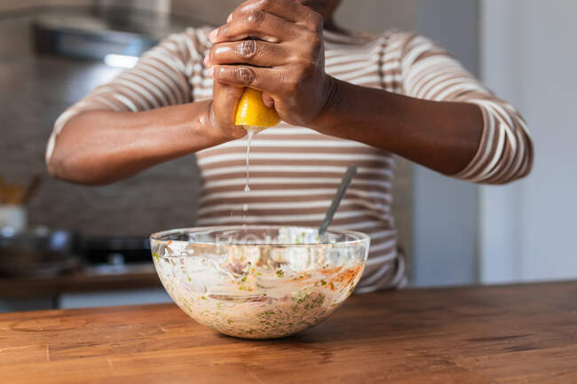 Cultivo irreconocible hembra étnica exprimiendo limón fresco sobre un tazón con comida en la mesa en la cocina - foto de stock