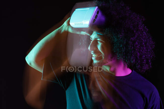 Delizioso maschio latino con capelli ricci e moderni occhiali di realtà virtuale in piedi in studio su sfondo nero con luci al neon — Foto stock