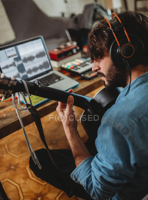 Back view of young man in headphones playing on guitar near table with laptop and synthesizer at home — Stock Photo