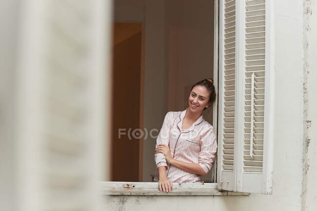 Mujer pacífica en pijama de pie en la ventana y mirando a la cámara en casa - foto de stock