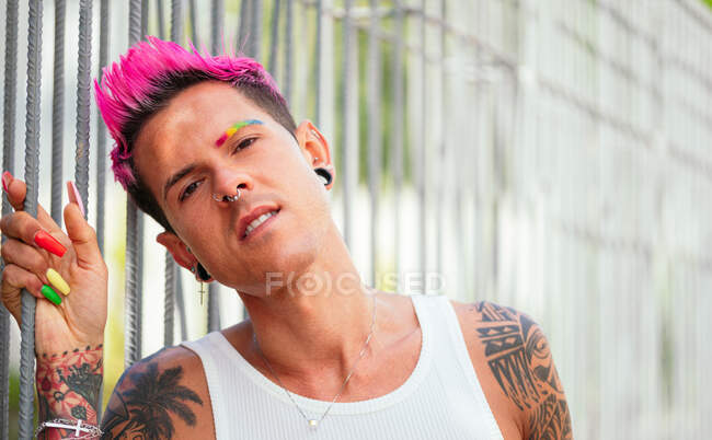Queer male with bright pink hair and colorful nails standing in street and leaning on metal fence while looking at camera — Stock Photo