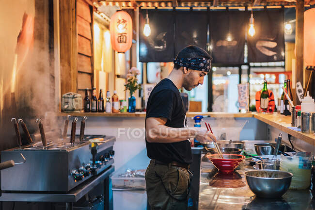 Side view of man in bandana standing at counter and cooking ramen in modern Asian cafe — Stock Photo