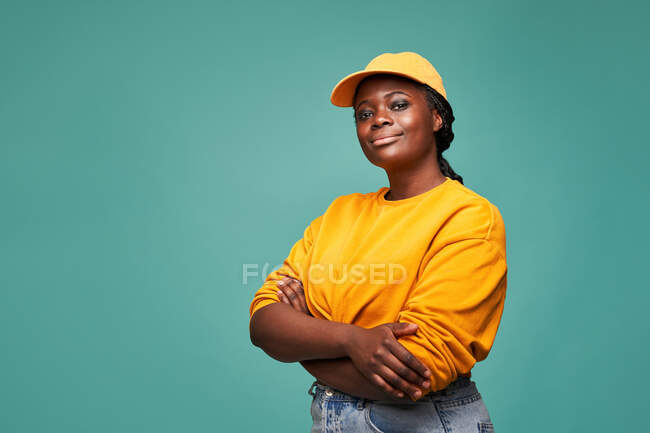 Happy plump African American female in yellow sweater and cap with arms crossed looking at camera while standing against blue wall — Stock Photo