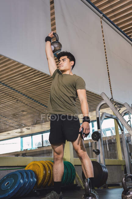 Hombre asiático entrenando hombros y brazos con pesadas pesas pesas en el gimnasio durante el entrenamiento funcional y mirando hacia otro lado - foto de stock