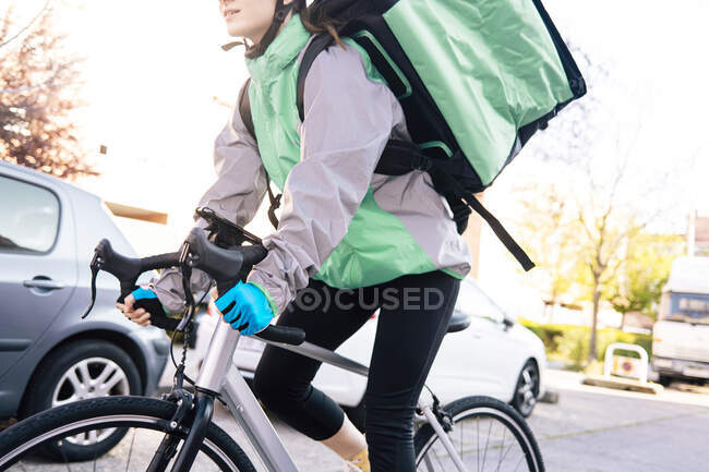 Low angle crop of female courier with thermal bag riding bike on street while making delivery on sunny day in city — Stock Photo