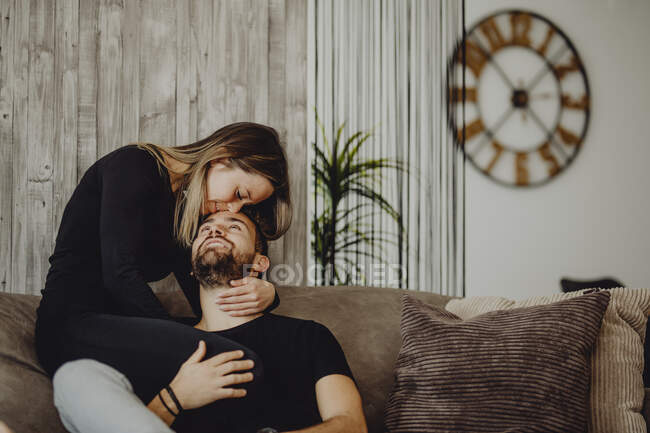 Усміхнена жінка обіймає і цілує веселого чоловіка в чоло, сидячи на зручному дивані вдома разом — стокове фото