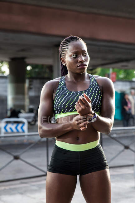 Афроамериканка держит руку на пульсе и смотрит в сторону во время тренировки на городской улице в солнечный день — стоковое фото