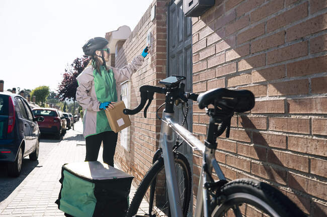 Female courier with parcel pressing button on intercom device on brick wall while making delivery on sunny day in town — Stock Photo