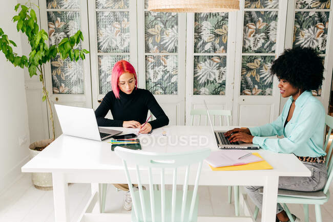 Концентрированный розовый волосатый фрилансер рисунок с карандашом и позитивной афроамериканской предпринимательницей, работающей с ноутбуком, сидя вместе за столом дома — стоковое фото