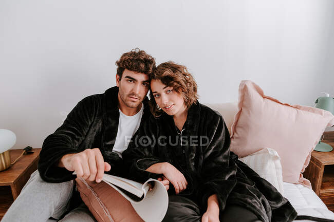 Zufriedenes Paar in Bademänteln entspannt auf der heimischen Couch, während es sich umarmt und in die Kamera blickt — Stockfoto