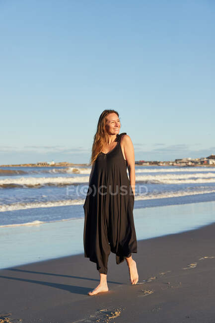 Sonriente hembra en vestido de verano de pie en la orilla del mar de arena y mirando a la cámara - foto de stock