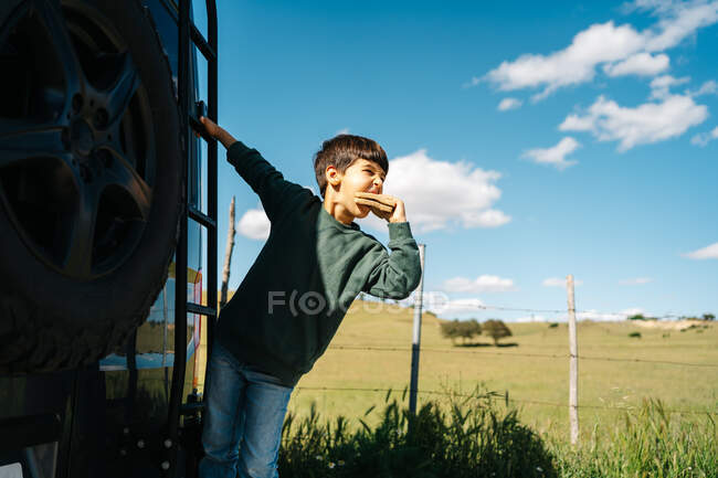 Carino piccolo viaggiatore mordere gustoso panino e distogliere lo sguardo mentre appeso sulla scala del moderno camper il giorno d'estate in campagna — Foto stock